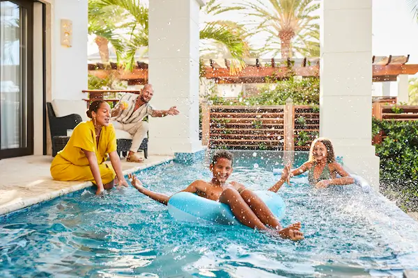 Family having fun by a pool, with two children splashing water and one child floating on an inflatable tube, while two adults watch and smile nearby, set in a tropical, resort-like environment.