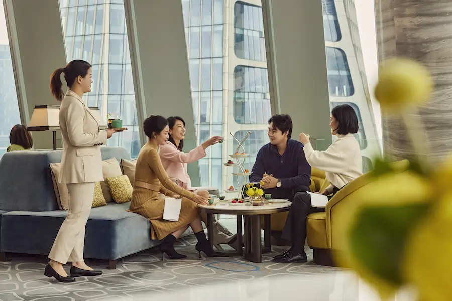A group of people enjoying an elegant afternoon tea in a modern lounge with large windows, high-rise buildings visible in the background, and a server attending to them, representing a sophisticated urban experience in Asia.
