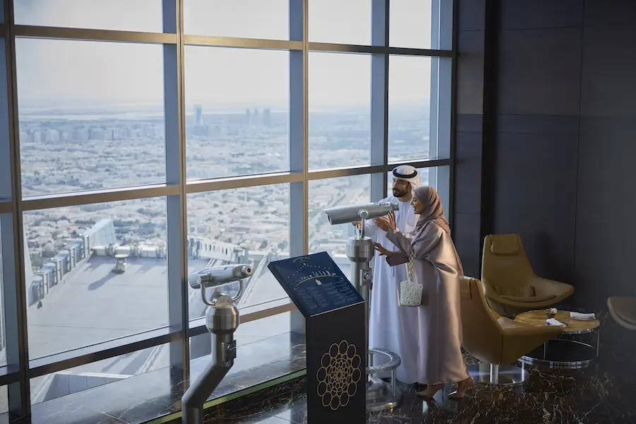 A couple dressed in traditional Middle Eastern attire using binoculars to view a cityscape from the observation deck of a modern high-rise building, symbolizing a scenic experience in the Middle East.