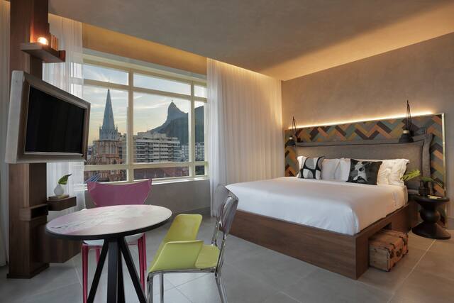 Guest room with bed, cafe table and chairs, TV, and large window with views of the city and mountains
