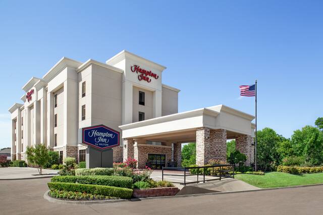 Exterior do hotel Hampton Inn de boas-vindas, com vegetação exuberante, porte cochere e céu azul.
